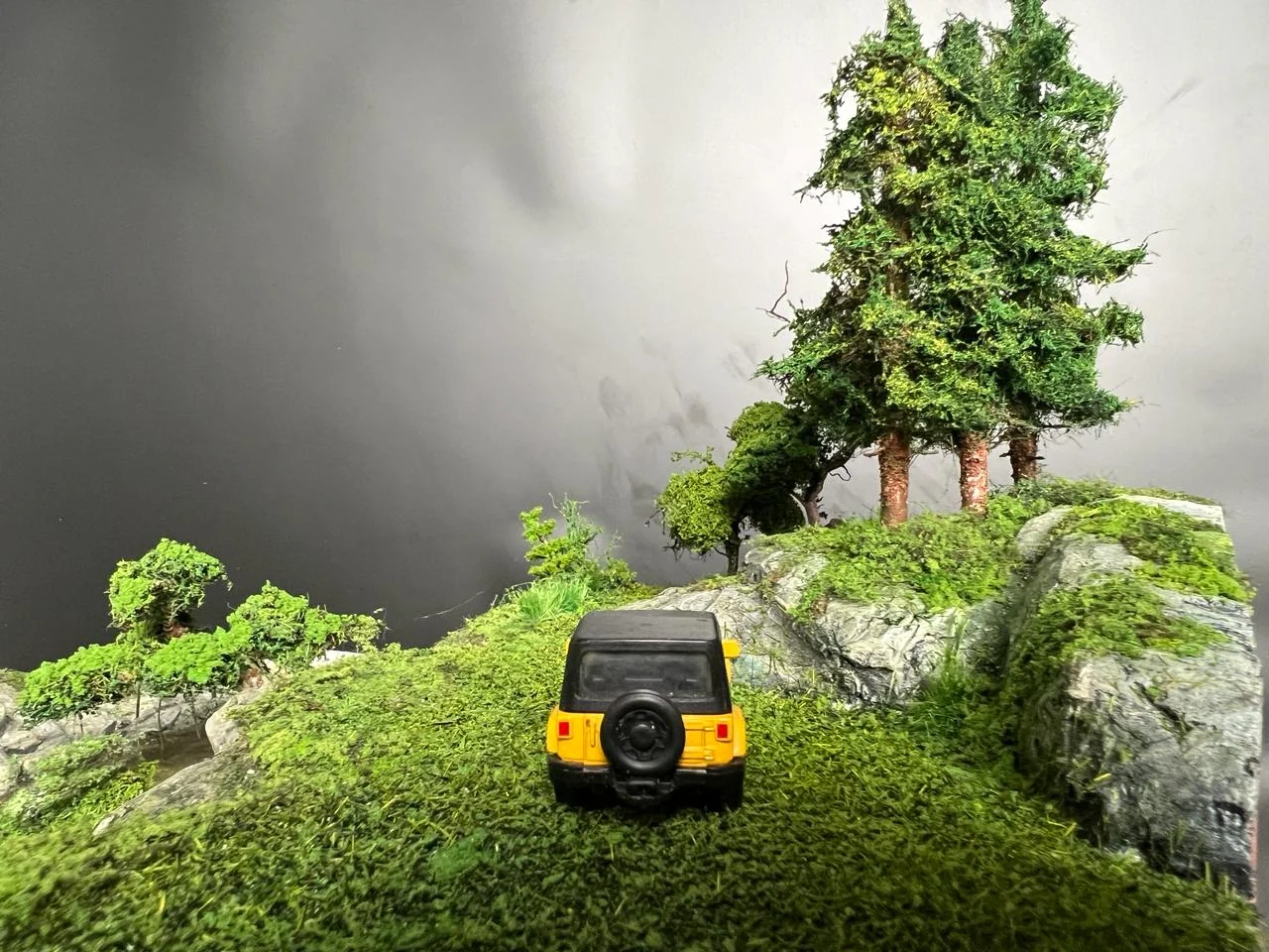 Miniature vehicle approaching pine trees in an English countryside diorama.