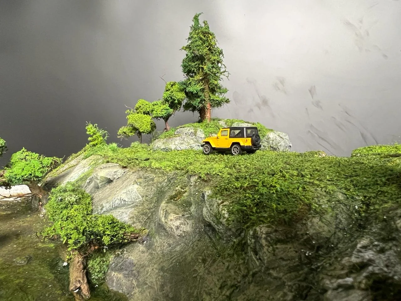 Yellow miniature vehicle on a green rocky ridge with detailed trees.