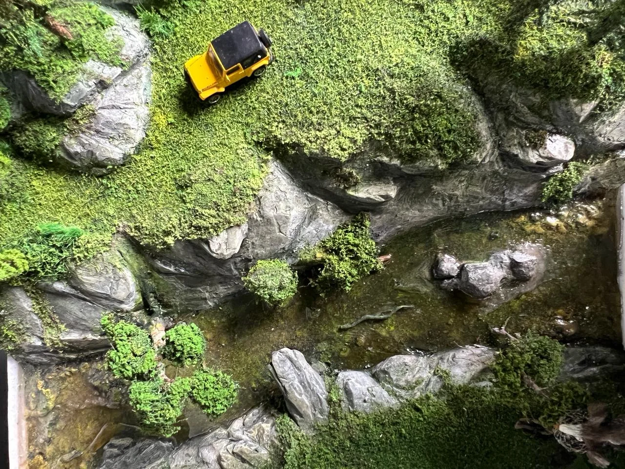 Topographic view of miniature terrain with stream, rocks, trees, and yellow vehicle.